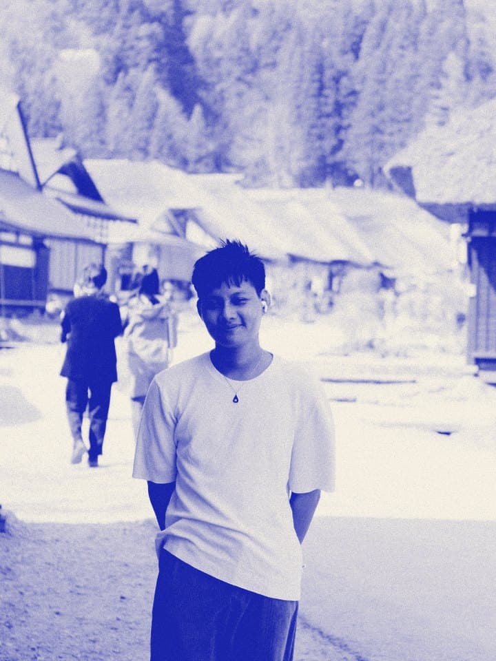 Din standing on a street of old thatched roof houses, hands behind his back